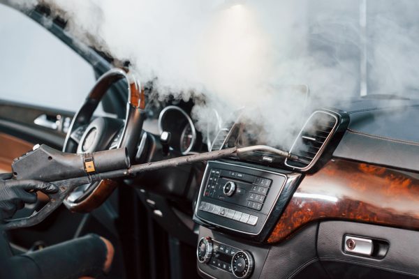 Modern black automobile get cleaned inside by steam equipment by worker inside of car wash station.