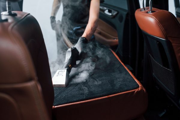 Uses steam cleaner. Modern black automobile is in service by woman inside of car wash station.